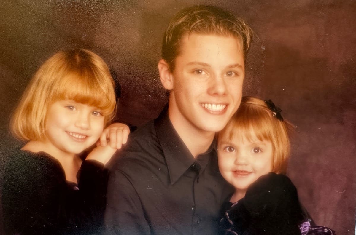 Three smiling children pose for a portrait against a brown backdrop, capturing a warm recollection. An older boy in a dark shirt sits in the middle, with a young girl and toddler girl in black velvet dresses on either side of him.