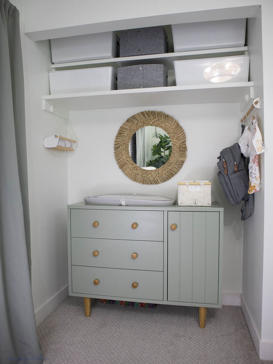 a view of the closet with dresser, storage bins and wall mounted shelves, mirror, and hanging rod in the baby nursery.