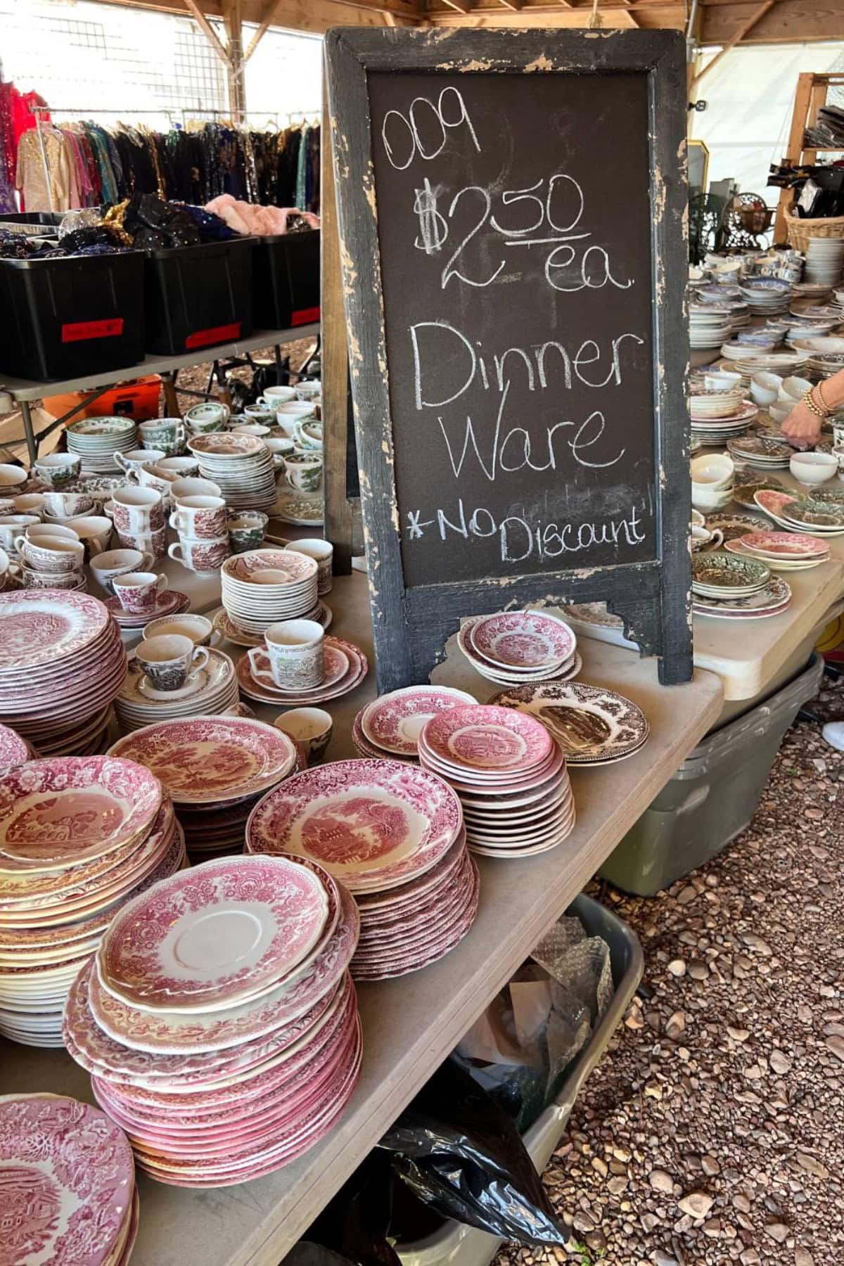 Stacks of assorted vintage dinnerware, including plates, bowls, and teacups, are displayed on tables at the Round Top outdoor market next to a chalkboard sign reading "$2.50 ea Dinner Ware *No Discount.