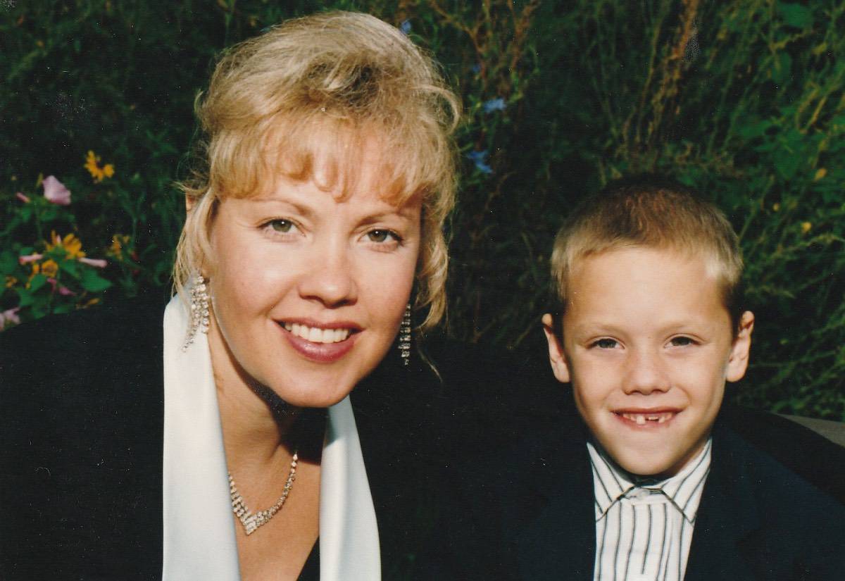 A smiling woman with blonde hair and a young boy in formal attire pose outdoors, surrounded by greenery and flowers, capturing a warm moment of recollection.