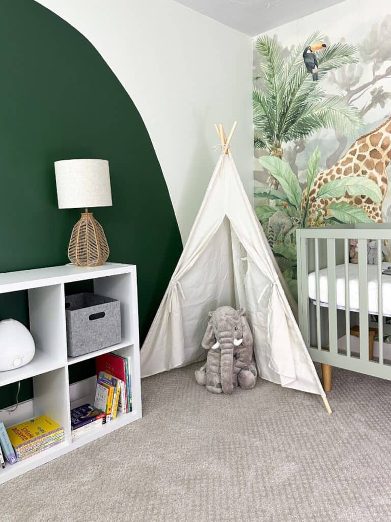 a corner of the room with a view of the bookshelve wall, toy tent, and mural wall with a crib