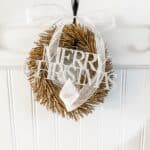 small gold wreath with a metal merry christms sign and a white ribbon bow handing in the front entrywy