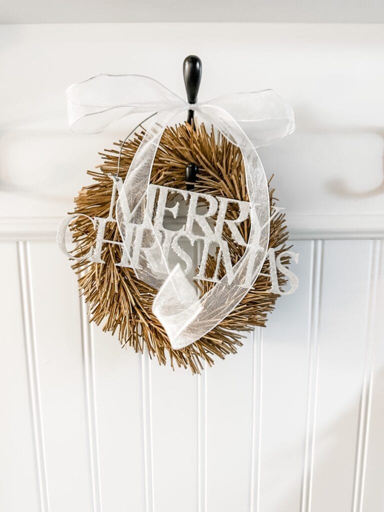 small gold wreath with a metal merry christms sign and a white ribbon bow handing in the front entrywy