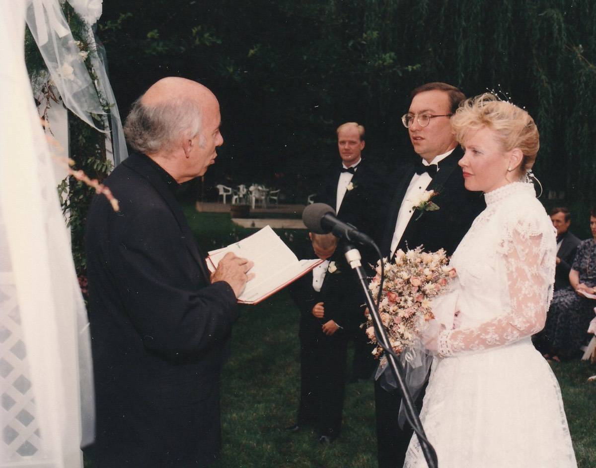 A bride and groom stand together outdoors facing an officiant holding a book, creating a beautiful recollection. The bride wears a white lace dress and holds a bouquet; the groom wears a tuxedo. Guests are seated in the background.