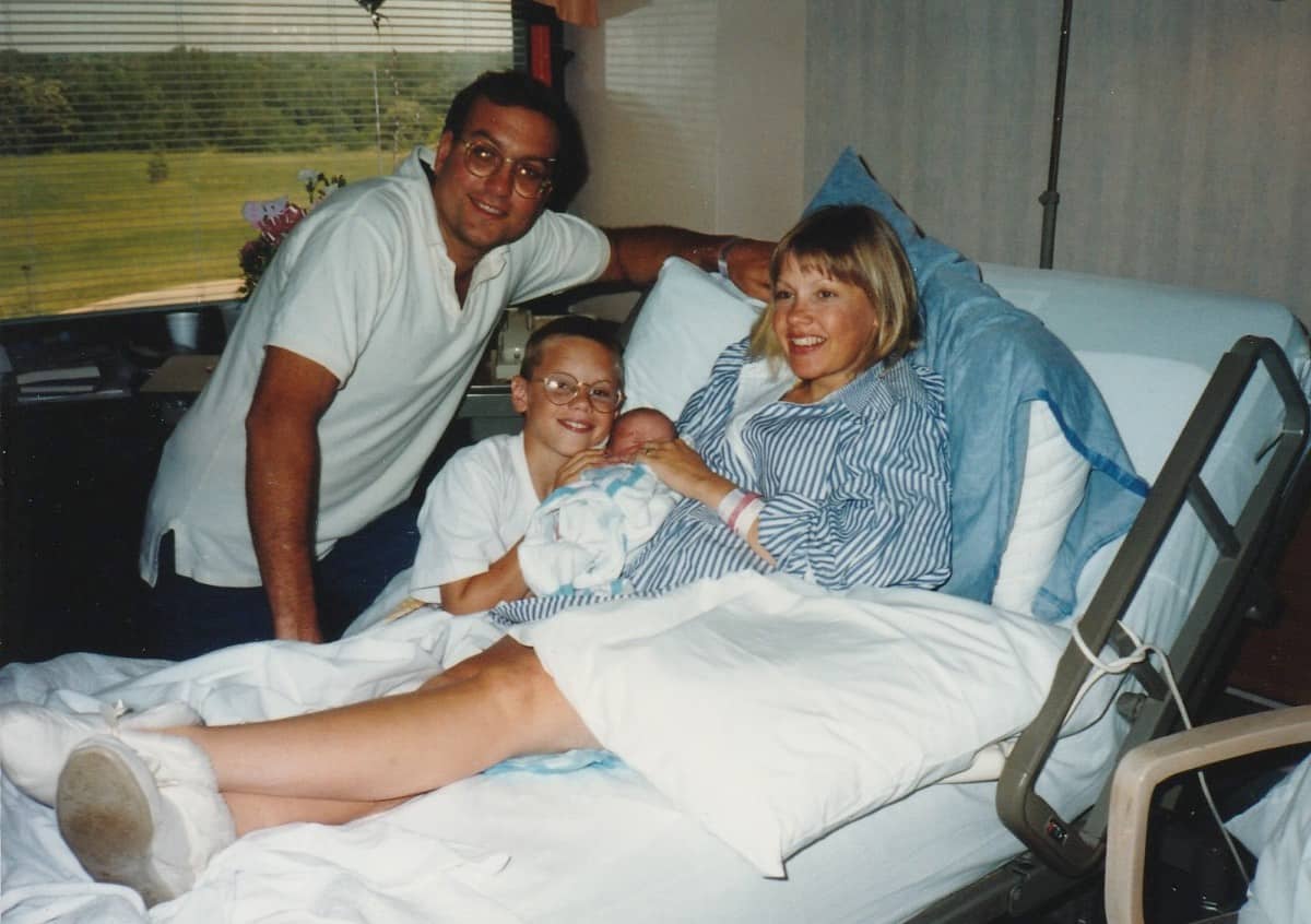 A woman lies in a hospital bed holding a newborn, with a smiling man beside her and a boy in glasses sitting nearby, all looking happy—a tender recollection framed by the greenery outside the window.