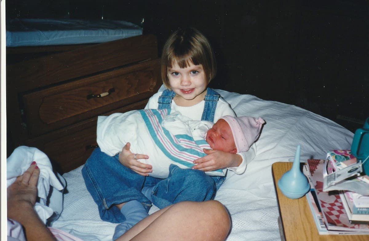 A young girl in overalls sits on a bed, smiling and holding a newborn baby wrapped in a blanket and wearing a pink hat—a tender moment that feels like a cherished recollection. An adult's hand and leg are partially visible in the foreground.