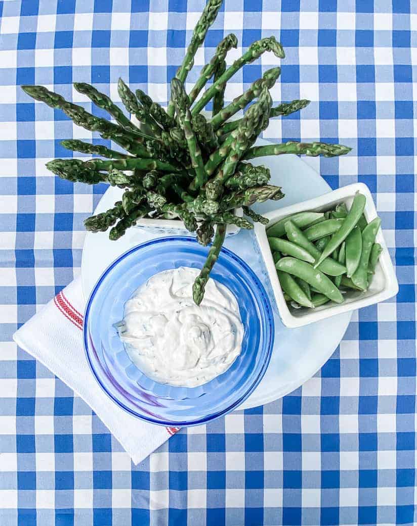 A plate with fresh asparagus stands upright in a blue glass, next to a bowl of snap peas, and a bowl of creamy dip on a blue and white checkered tablecloth. A folded white napkin with a red stripe is placed beside the plate.