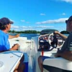 Two men sit on a boat, one steering and the other pointing toward the water—a scene filled with recollection. The sky is blue with scattered clouds, trees line the shore, and it’s a sunny, relaxing day on the water.