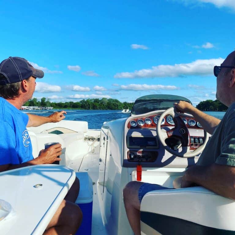 Two men sit on a boat, one steering and the other pointing toward the water—a scene filled with recollection. The sky is blue with scattered clouds, trees line the shore, and it’s a sunny, relaxing day on the water.