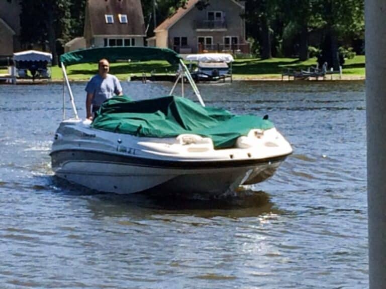 A person is driving a white motorboat with a green canopy and boat cover on a lake, with houses and trees lining the shoreline in the background—a scene perfect for recollection.