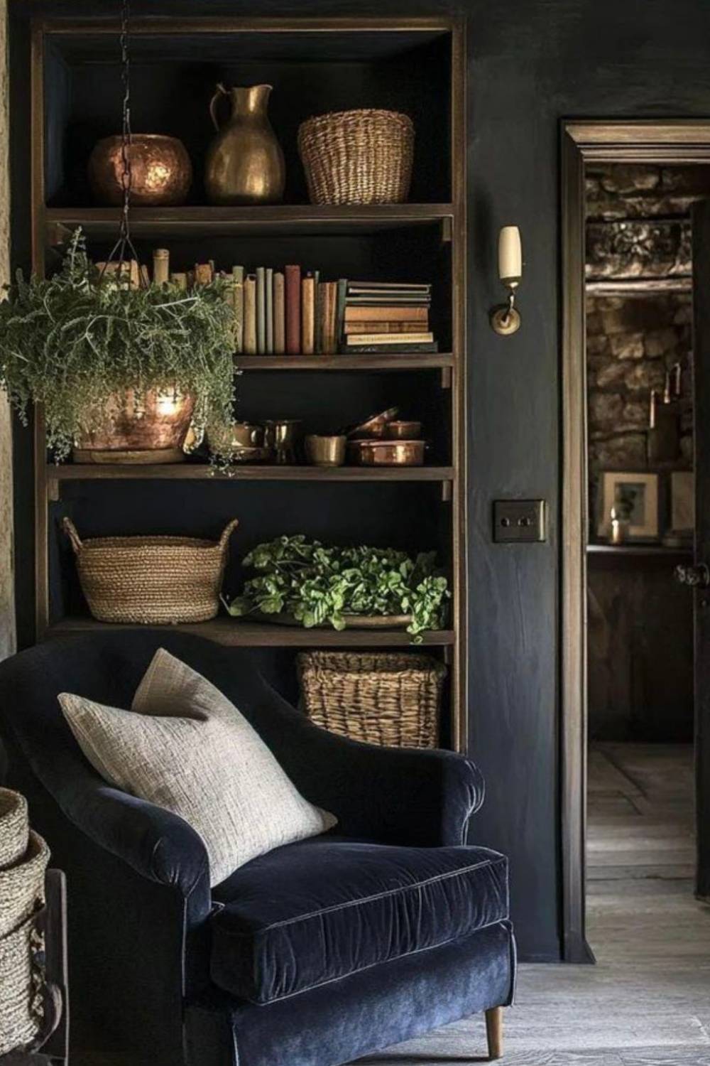 A cozy reading nook with a dark velvet armchair and light pillow showcases dark and moody interior design, nestled beside bookshelves filled with books, baskets, copper pots, and green plants. A hanging plant and a doorway to a rustic stone-walled room complete the scene.