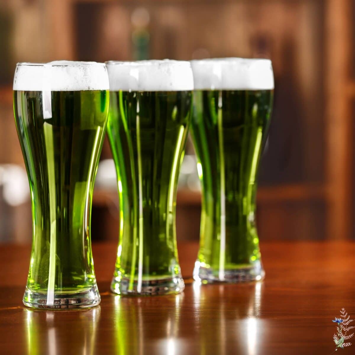 Three tall glasses filled with green beer, topped with white foam, are placed on a polished wooden surface, creating a festive St. Patrick's Day tablescape against a blurred background.