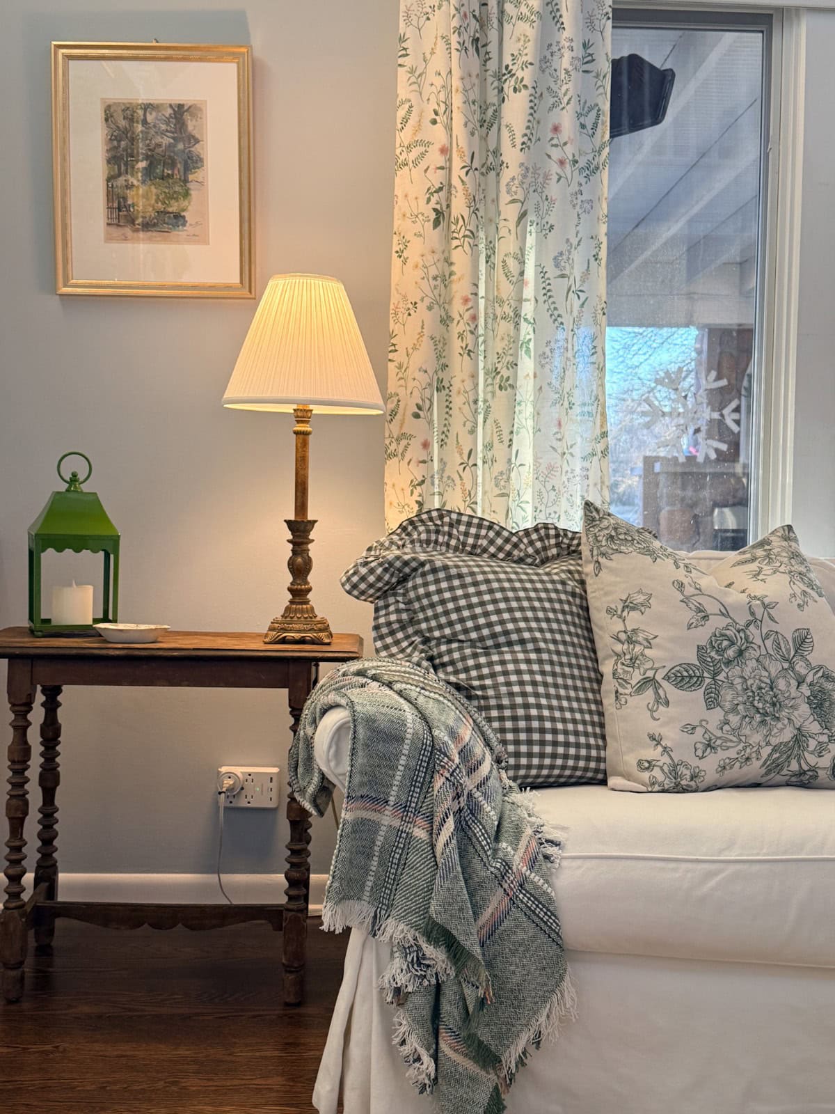I changed my living room into a cozy corner with a white sofa, patterned pillows, and a plaid throw. A wooden side table holds a lamp, green lantern, and framed picture against a light blue wall. A floral curtain hangs by the window.
