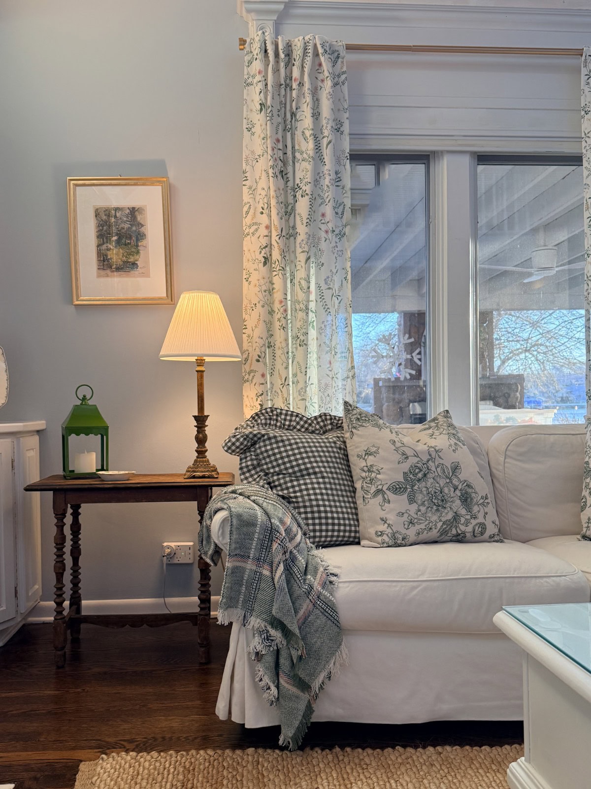 A cozy living room corner where I changed my living room with a white sofa decorated with patterned pillows and a plaid throw, a wooden side table holding a lamp and green lantern, floral curtains, and a large window letting in natural light.