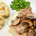 A plate featuring savory meatloaf topped with sautéed mushrooms, accompanied by creamy mashed potatoes and a side of vibrant peas.