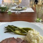 A table setting showcases a delicious meal with sliced beef, creamy mashed potatoes, and asparagus drizzled with sauce on a pristine white plate. In the background, wine glasses and another softened plate complement the cozy home decor elements enveloping the scene.