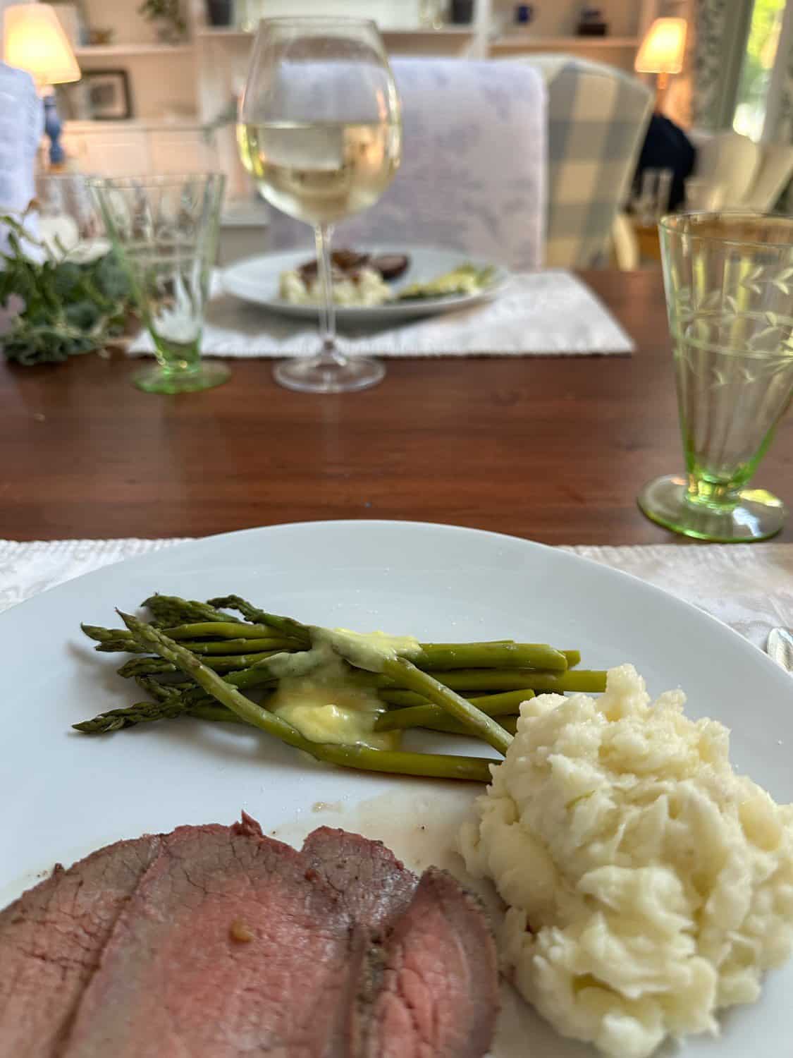 A table setting showcases a delicious meal with sliced beef, creamy mashed potatoes, and asparagus drizzled with sauce on a pristine white plate. In the background, wine glasses and another softened plate complement the cozy home decor elements enveloping the scene.