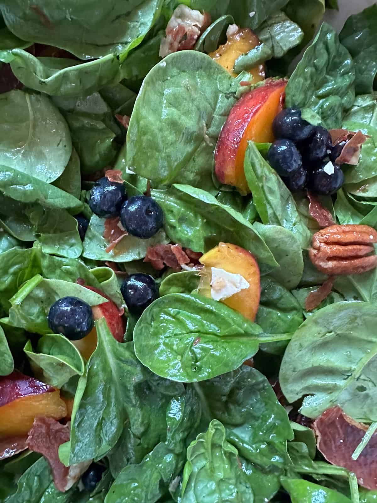 A close-up of a vibrant spinach salad featuring sliced peaches, blueberries, pecans, and bits of crispy prosciutto. The fresh ingredients are mixed together and lightly coated in dressing, creating a colorful masterpiece.