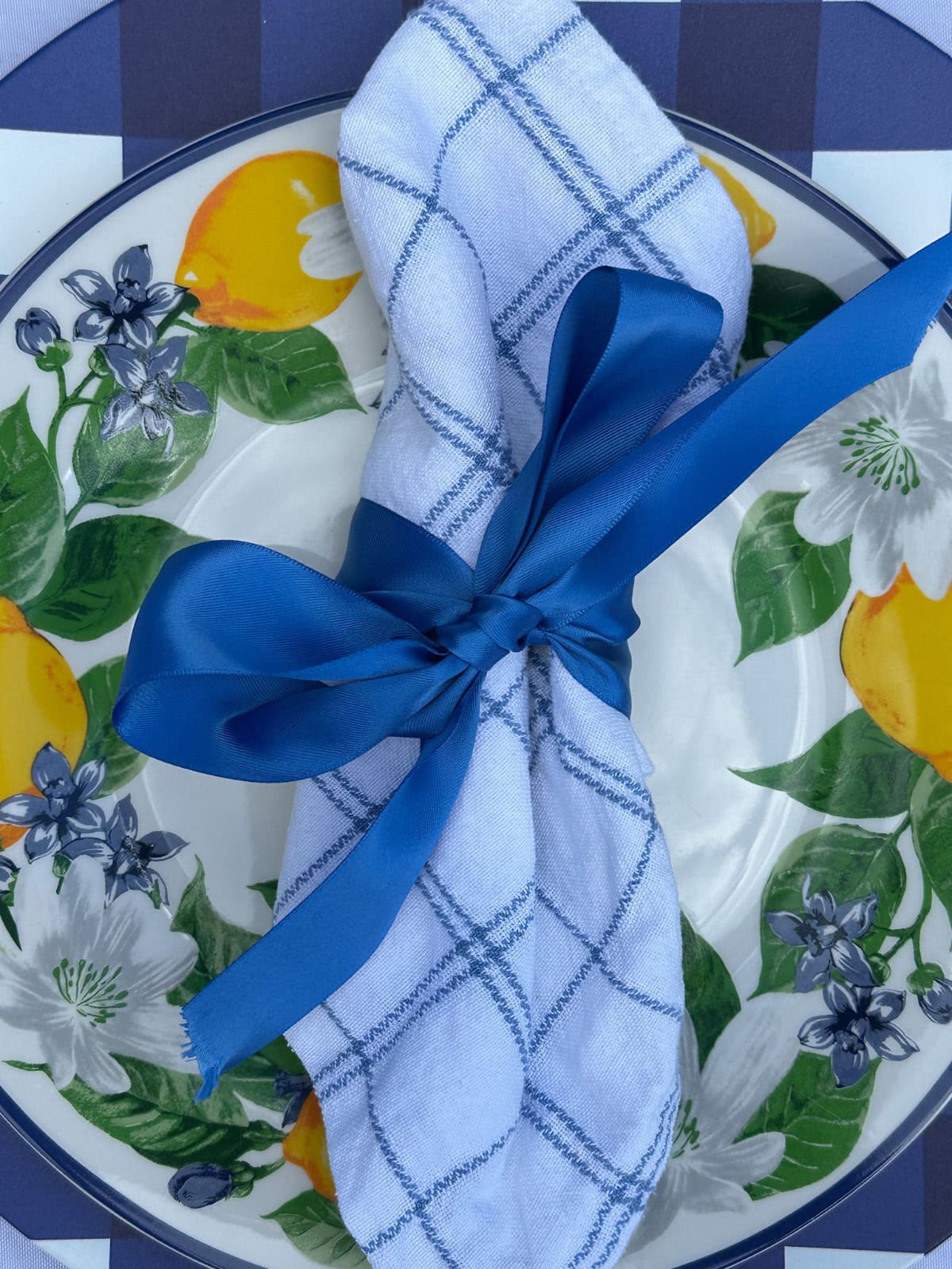 A white napkin with blue stripes, folded and tied with a blue ribbon, is placed on a white plate with a decorative pattern of green leaves, white flowers, and yellow lemons. The plate sits on a blue and white table setting.