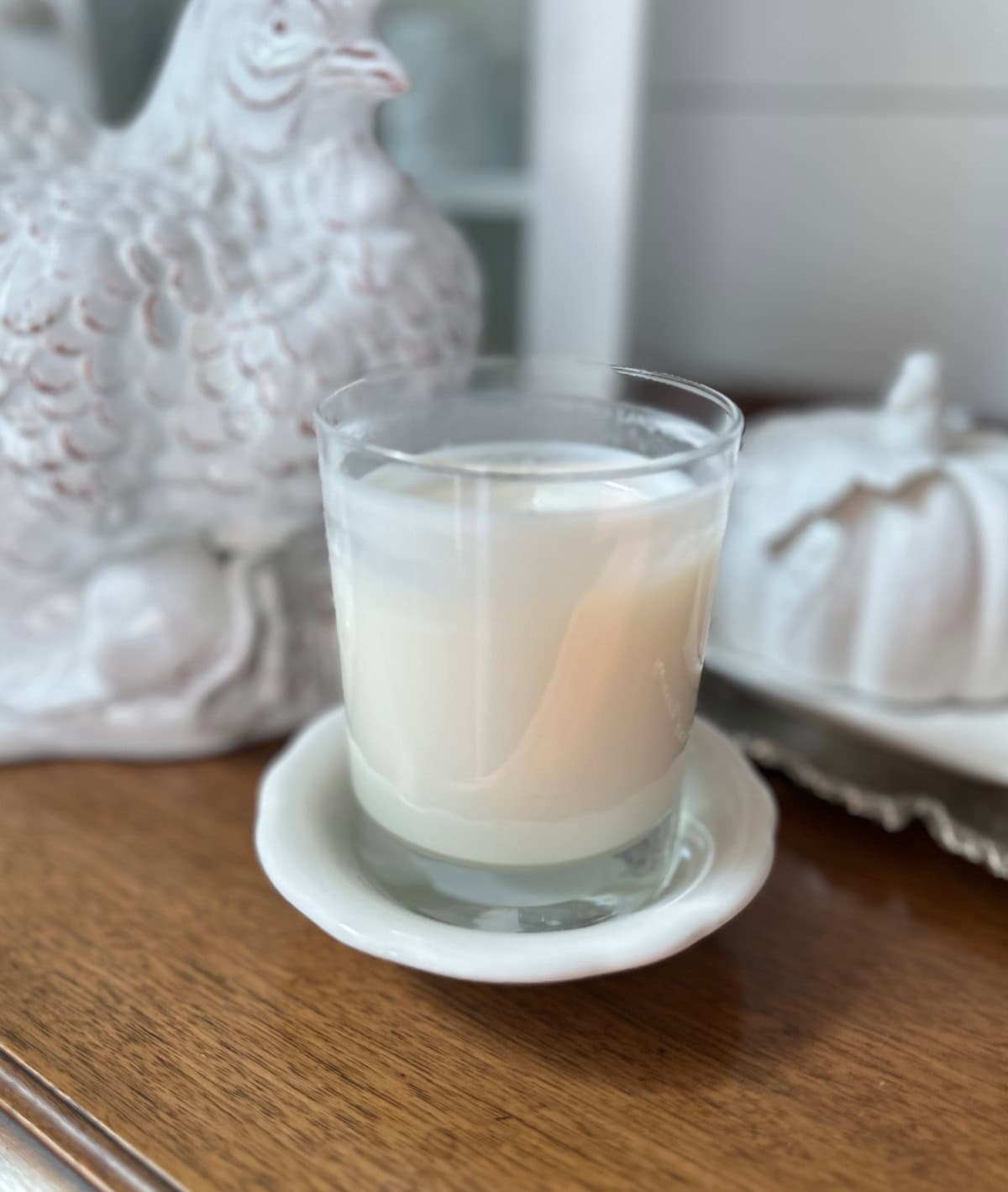 A white candle in a glass holder sits on a small white saucer. The candle and saucer are placed on a wooden surface. In the background, there is a white ceramic hen and a white pumpkin decoration, reminiscent of finds from Saturday shopping trips. The scene has a light and cozy aesthetic.