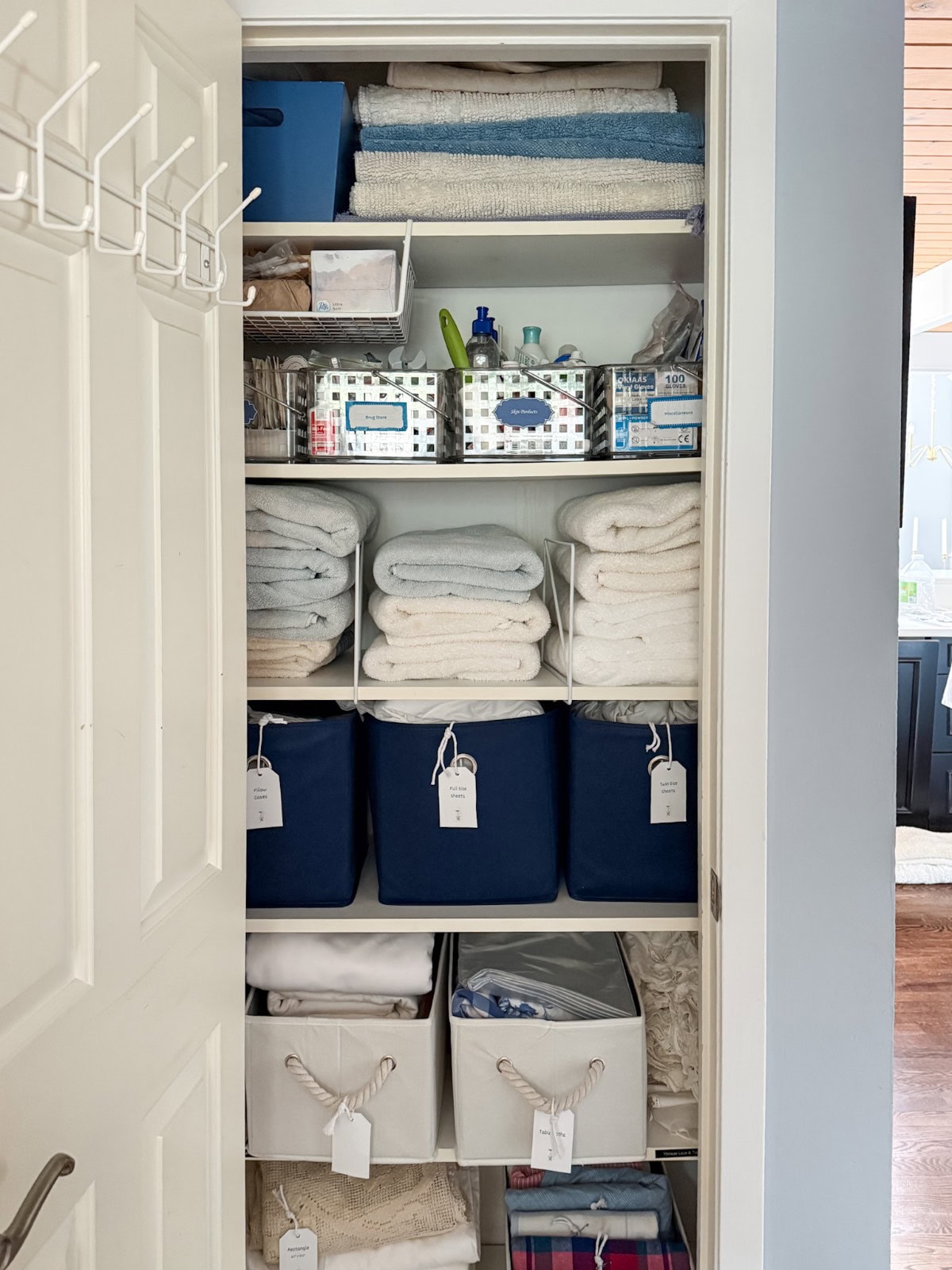 A neatly organized linen closet with shelves holding folded towels, labeled blue and white storage bins, and clear containers with toiletries offers great Organizing Tips for Bedrooms and Bathrooms. The closet is tidy and sorted, with tags on each bin and item.