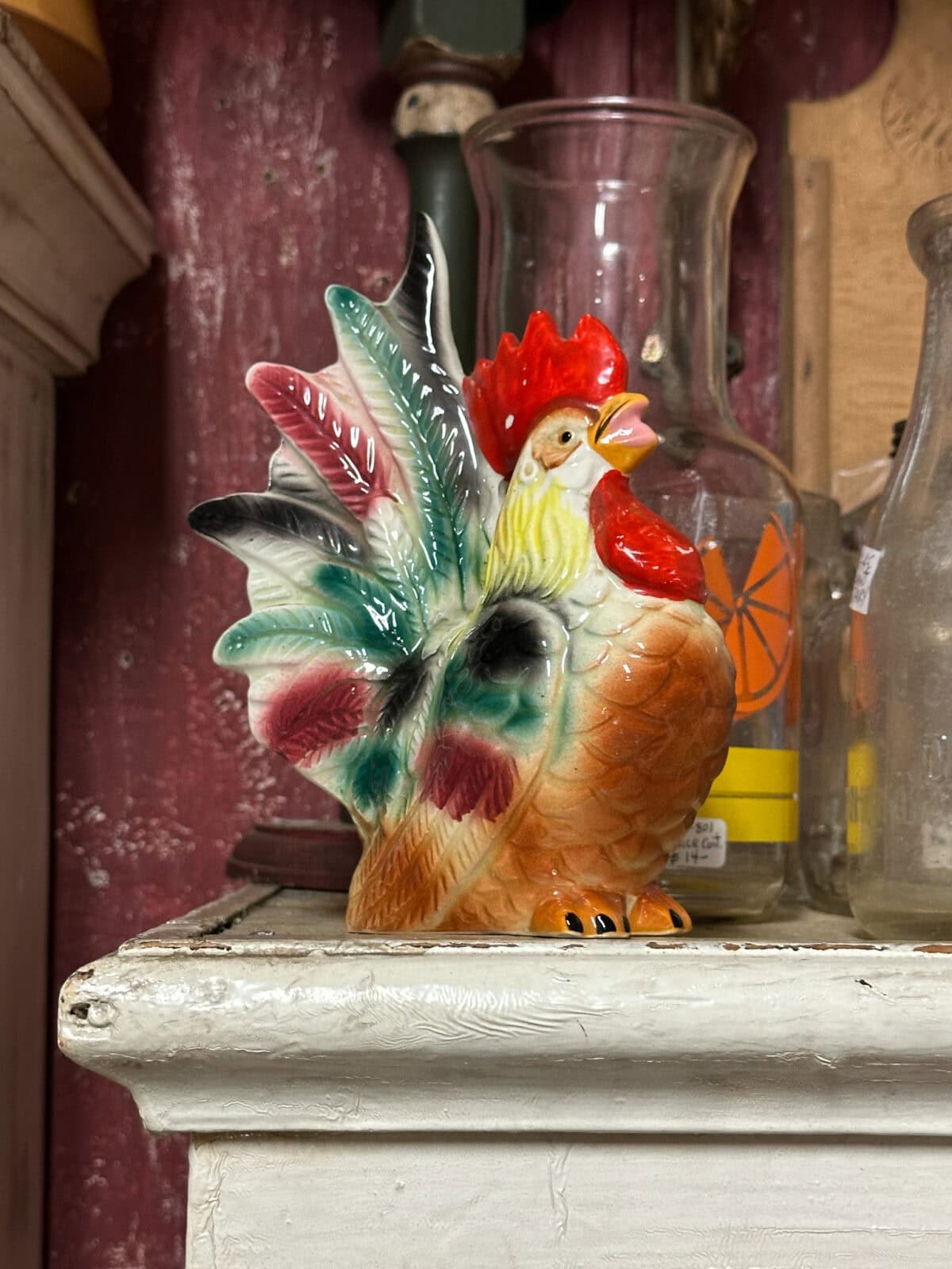 A colorful ceramic rooster figurine with vibrant feathers stands on a white wooden shelf. Behind it are glass bottles, one featuring an orange slice design. The reddish-brown hue of the wall enhances the rustic ambiance, making it perfect fall decor for your home.