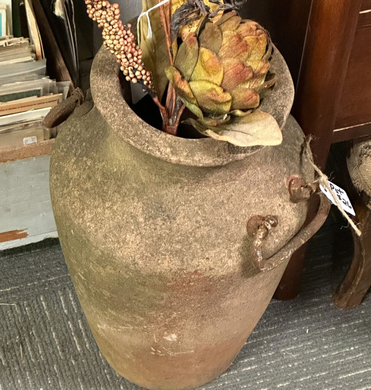 A large, weathered ceramic urn with two handles, containing dried floral arrangements, including an artichoke and berries, exemplifies fall decor. It is placed on a gray and white patterned carpet next to a wooden piece of furniture. A crate with papers is partially visible in the background.
