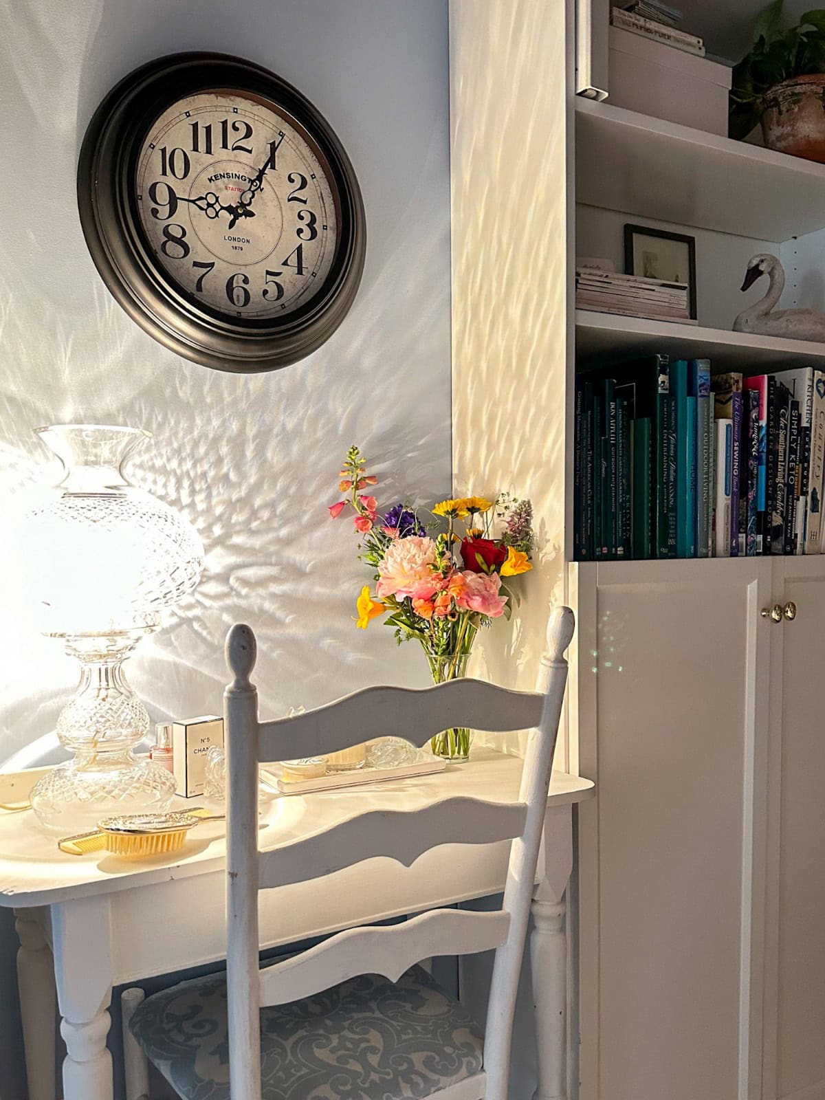 A cozy reading nook perfect for organizing living space, with a white chair, a table holding a glass lamp and colorful bouquet, a clock showing 7:50, and bookshelves neatly arranged with books and decorative items as sunlight casts patterns on the wall.