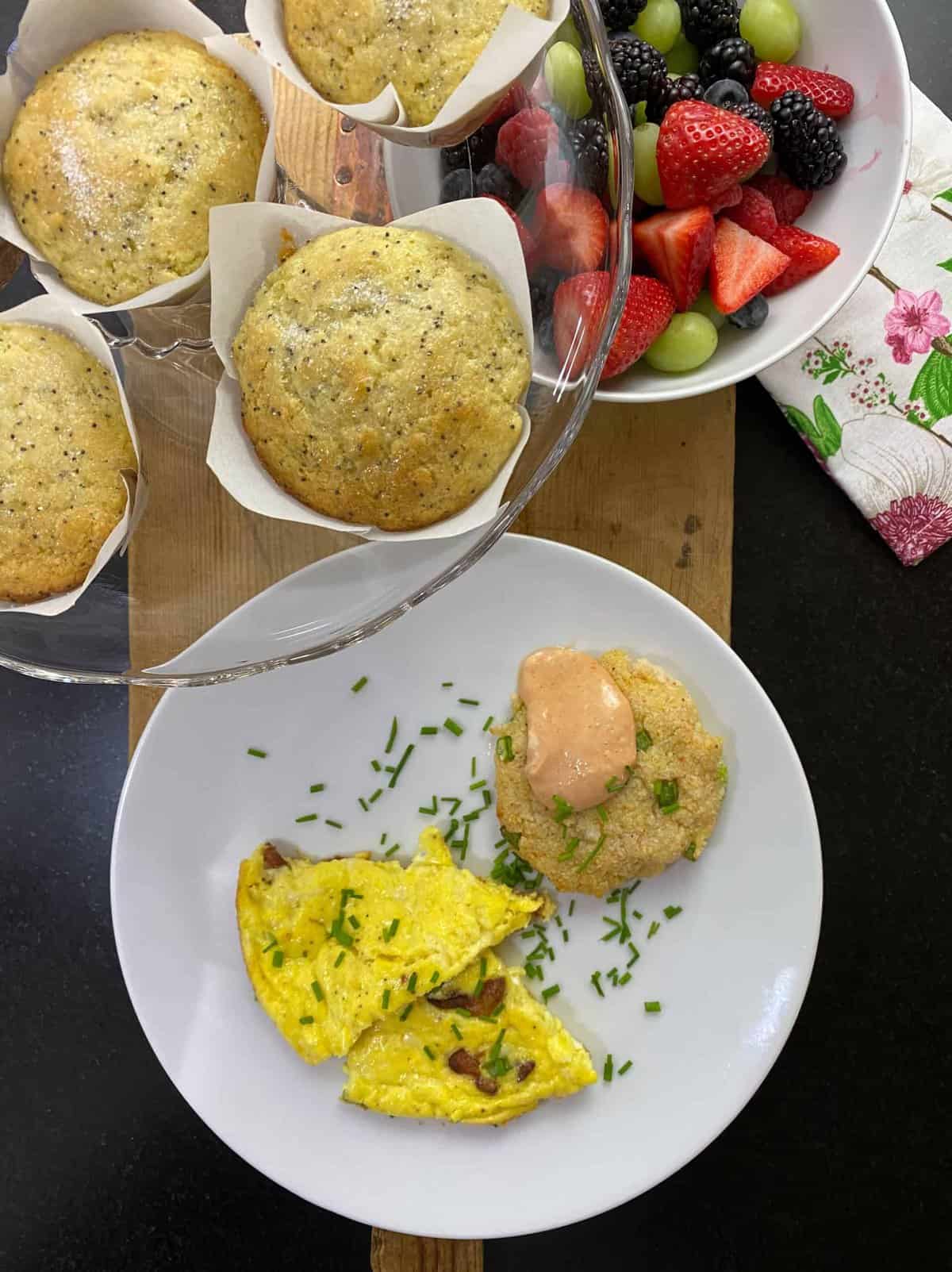 A breakfast scene with quinoa patty topped with sauce and chives on a plate, next to a split vegetable frittata. A glass tray holds lemon poppy seed muffins above, and a bowl of fresh berries and grapes is nearby. A floral napkin adds a decorative touch.