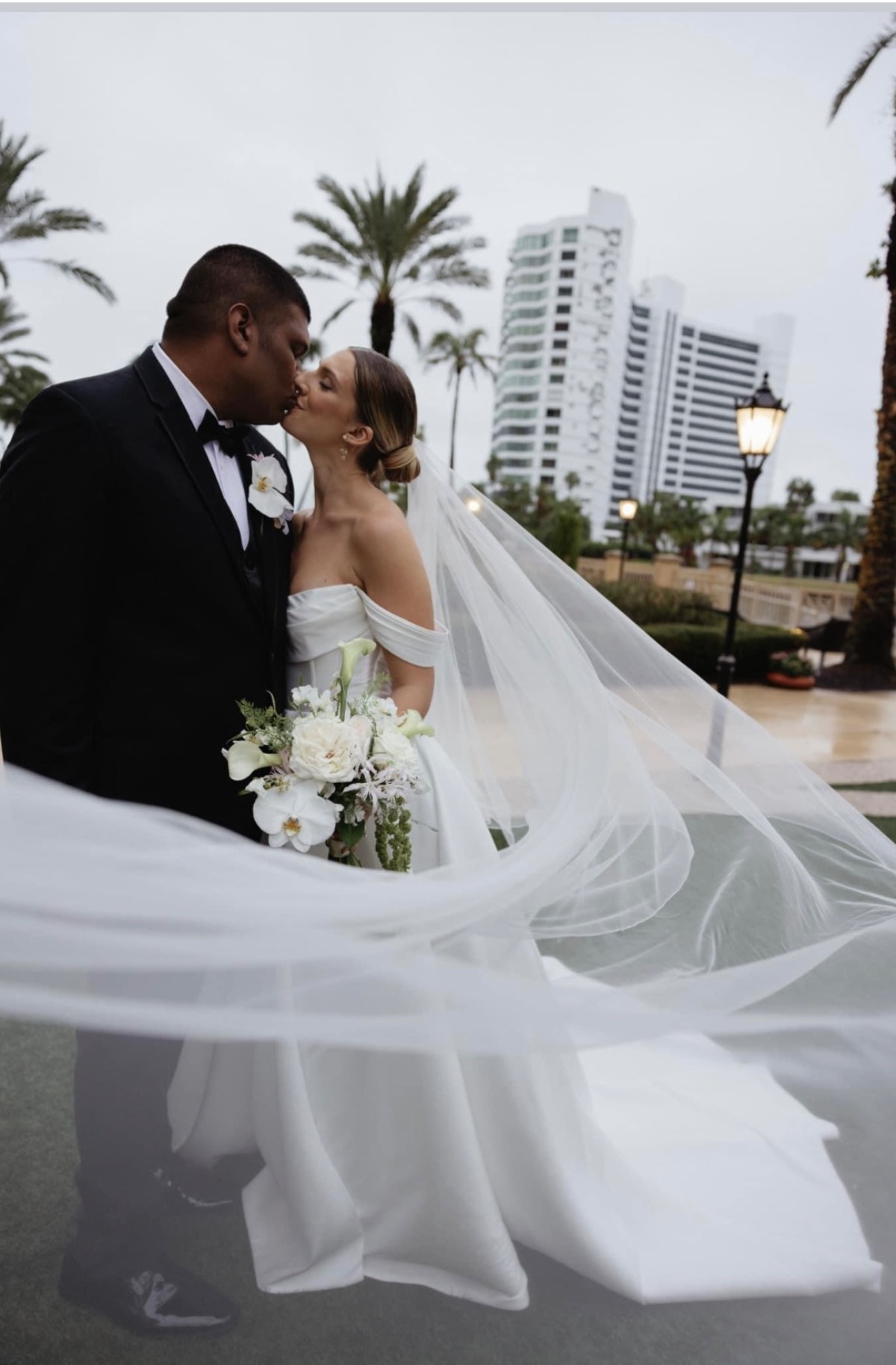 A couple shares a kiss on their wedding day, a moment of sweet recollection. The bride, adorned in a white gown with a long veil, holds a bouquet as the groom wears a black suit with a white boutonniere. In the background stand palm trees and a modern building.