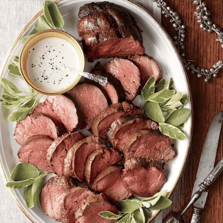 Sliced beef tenderloin arranged on an oval platter with fresh sage leaves. A small bowl of creamy white sauce with a spoon rests atop the meat. Silverware is placed beside the platter on a wooden surface.