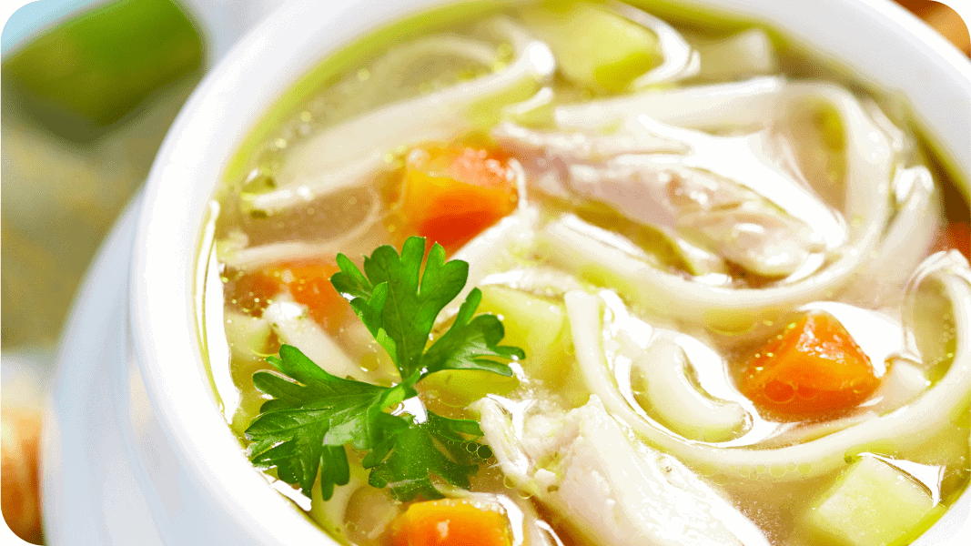 A close-up of a bowl of chicken noodle soup with visible noodles, chunks of chicken, carrots, celery, and a sprig of parsley on top, placed on a smooth white surface.