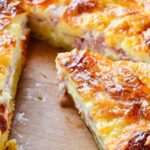 A close-up of a freshly baked quiche on a wooden cutting board. The golden-brown crust and cheesy surface are visible, with a slice partially removed, revealing layers of ham and creamy filling.