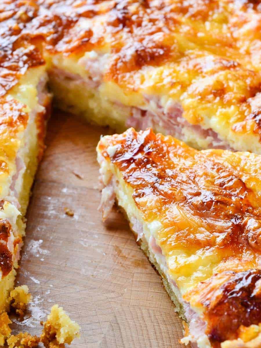 A close-up of a freshly baked quiche on a wooden cutting board. The golden-brown crust and cheesy surface are visible, with a slice partially removed, revealing layers of ham and creamy filling.