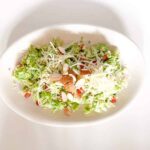A white oval plate containing a fresh salad with shredded Brussels sprouts, sliced almonds, grated cheese, and pomegranate seeds, set on a white background.