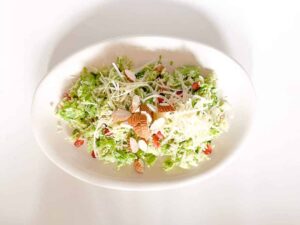 A white oval plate containing a fresh salad with shredded Brussels sprouts, sliced almonds, grated cheese, and pomegranate seeds, set on a white background.