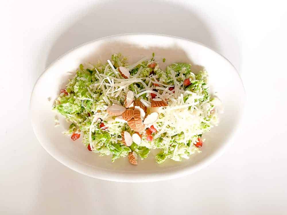 A white oval plate containing a fresh salad with shredded Brussels sprouts, sliced almonds, grated cheese, and pomegranate seeds, set on a white background.
