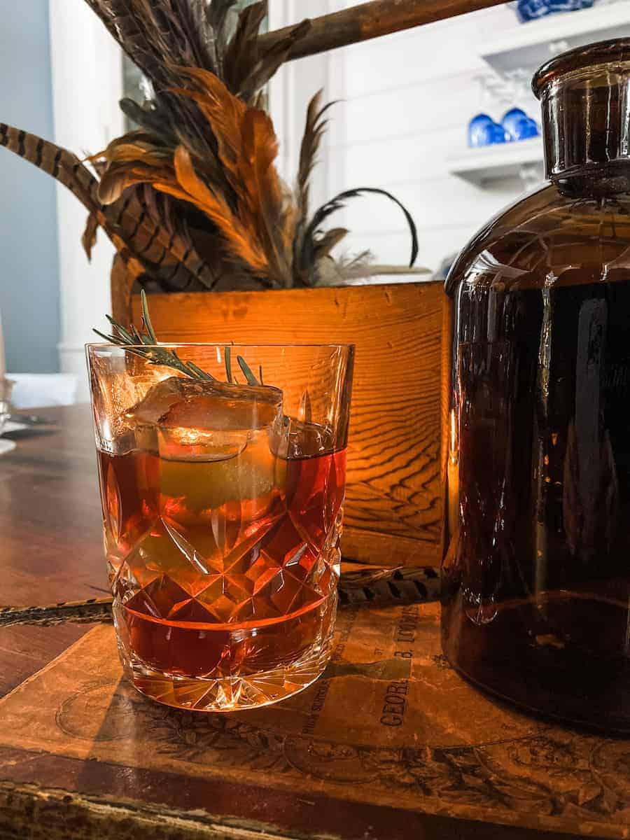 A crystal glass filled with an amber-colored cocktail, garnished with rosemary, sits on a wooden surface. A brown bottle and a rustic arrangement of feathers and a wooden container are in the background, giving a warm, cozy ambiance.