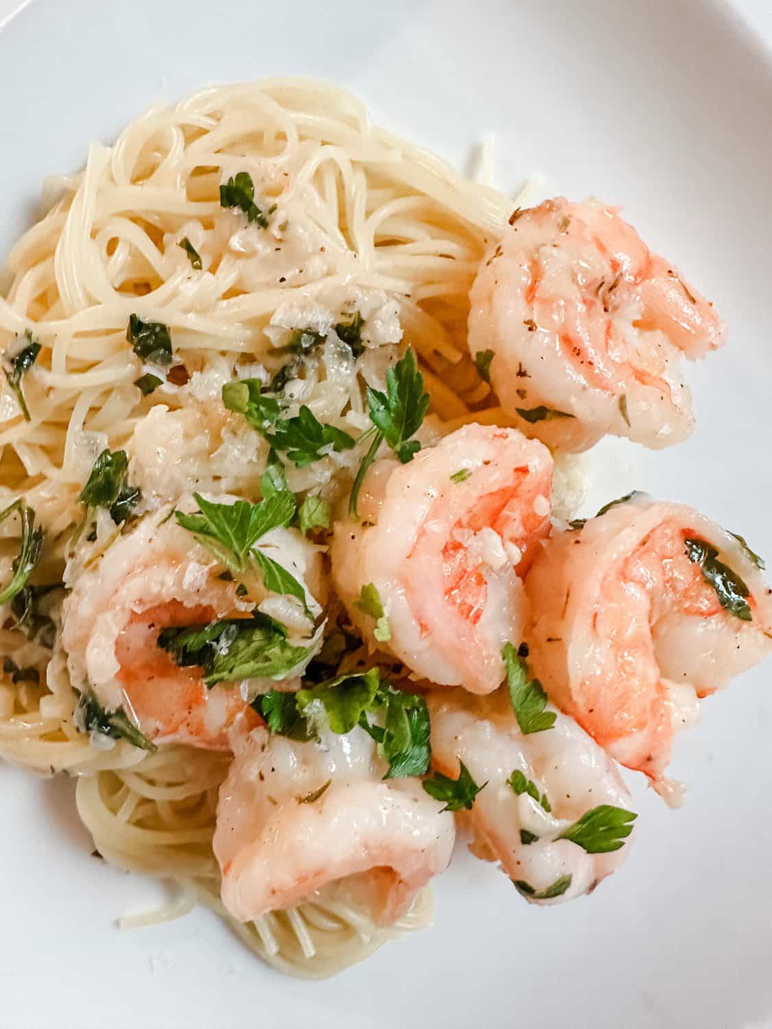 A plate of shrimp scampi with angel hair pasta, garnished with fresh parsley. The shrimp are pink and seasoned, nestled on lightly sauced pasta. The dish is served on a white plate.