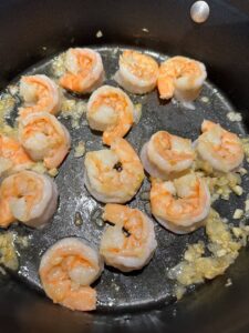 Shrimp cooking in a skillet with chopped garlic, turning pink as they heat. The shrimp are arranged in an even layer on the pan surface, surrounded by sizzling bits of garlic.