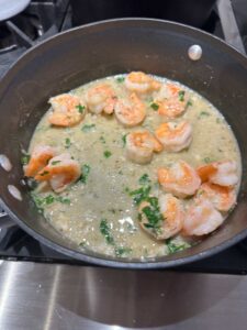 A skillet with cooked shrimp in a creamy, herb-speckled sauce on a stove. The shrimp are pink and nestled in a mix of green herbs and a thick, pale-colored sauce.