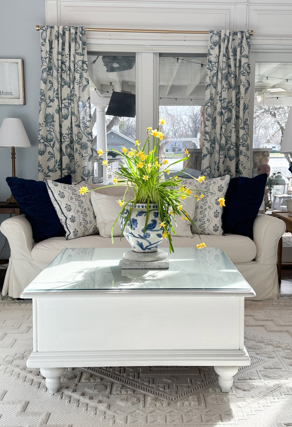 A cozy living room exudes an upscale look with its white sofa adorned with patterned blue and white pillows. A glass-topped coffee table holds a matching vase with yellow flowers. Large windows, dressed in floral curtains, allow natural light to elegantly fill the space.