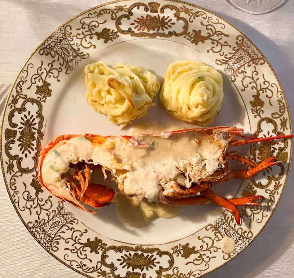 A decorative plate features a lobster halved with creamy sauce, accompanied by two portions of mashed potatoes piped in a spiral shape, set on a white tablecloth.
