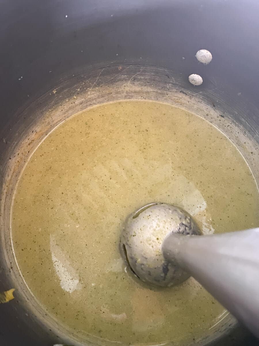 A blending immersion blender is placed in a pot with green soup being blended. The soup appears to have a creamy texture and remains slightly unblended. The pot's sides are splattered with bits of the soup mixture.