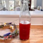 A glass bottle filled with red liquid stands on a wooden countertop. Beside it, a decorative wreath made of twigs and small flowers rests. The background features a well-lit room with windows and a chandelier.
