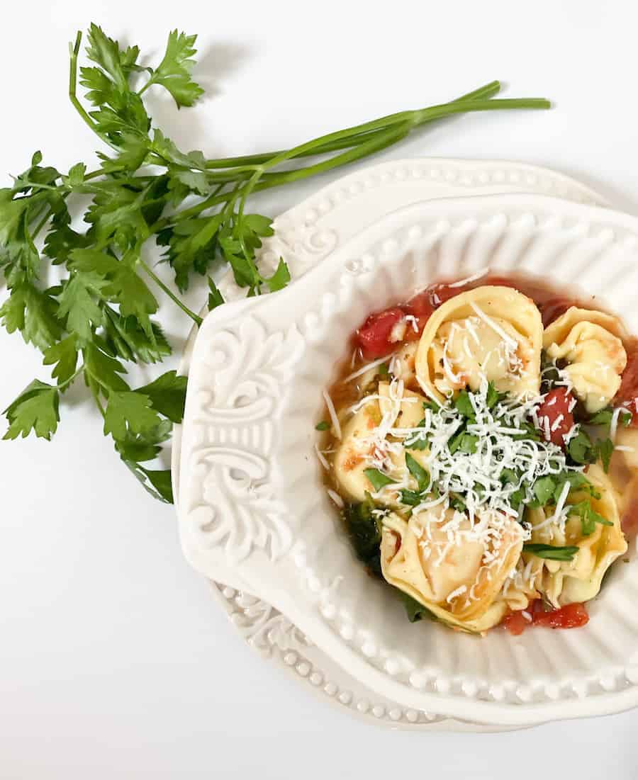 A decorative white bowl filled with tortellini pasta topped with grated cheese, tomato sauce, and fresh herbs. A sprig of parsley is placed beside the bowl on a white background.