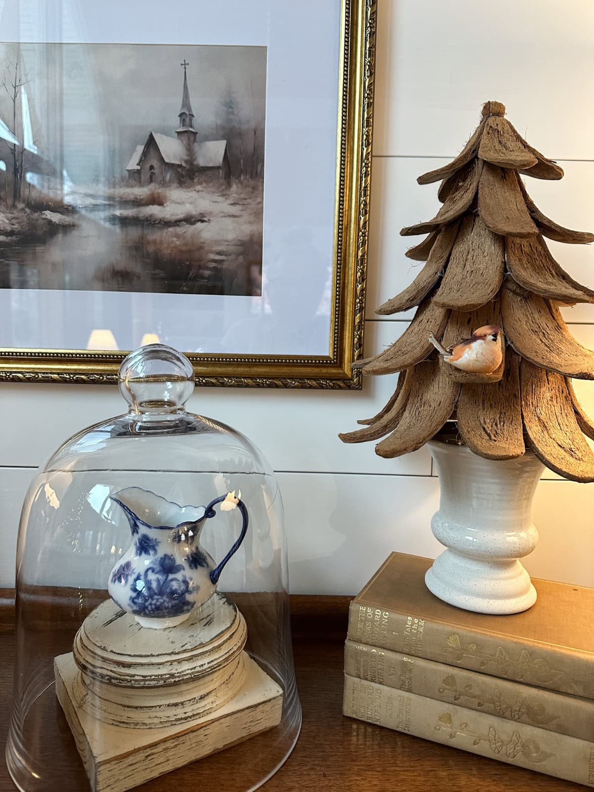 A cozy interior features a rustic wooden tree sculpture with an Old World Christmas charm, as a small bird figurine nestles on it. Nearby, a blue and white teapot under a glass dome sits gracefully on a wooden stand. A framed painting of a winter church scene hangs on the wall, completing the festive ambiance.