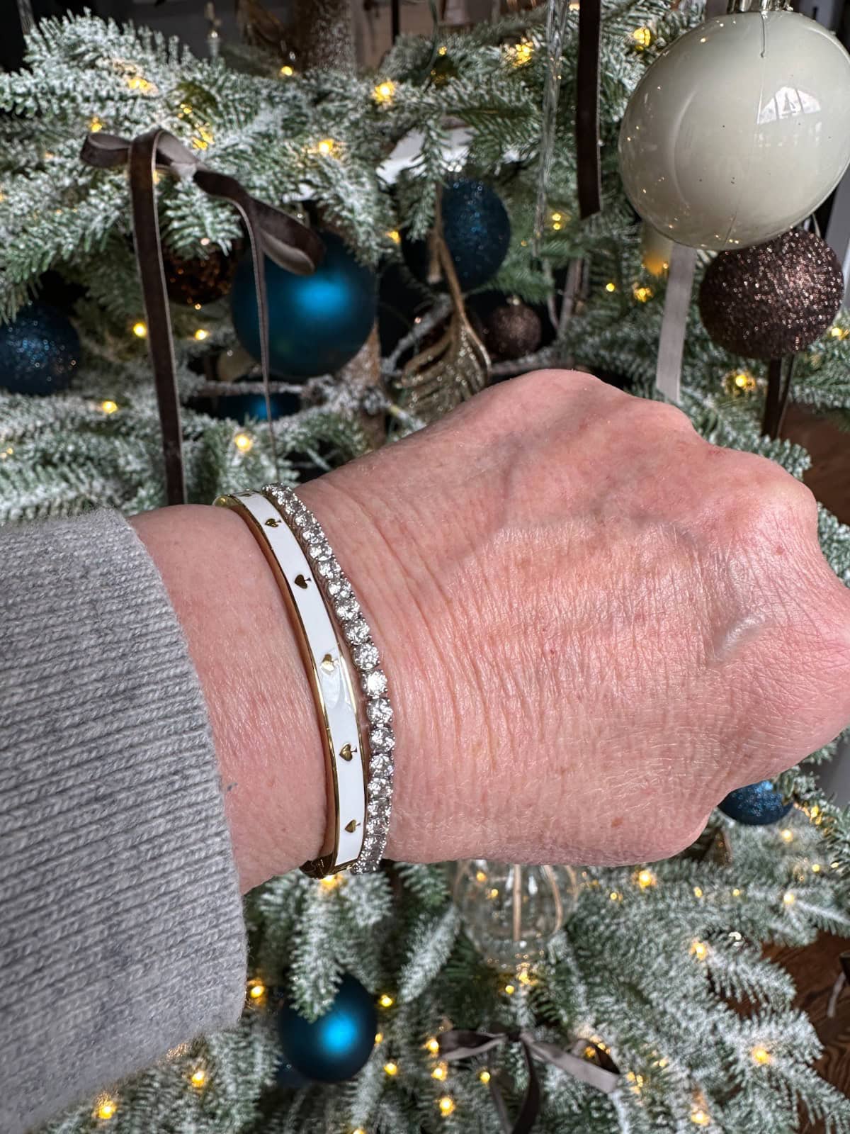 Favorite-Things | Living Large In A Small House, LLC A hand wearing a white and gold bracelet with heart-shaped studs and a sparkly diamond bracelet is held in front of a decorated Christmas tree, ready for Saturday Shopping among blue, white, and brown ornaments and twinkling lights.