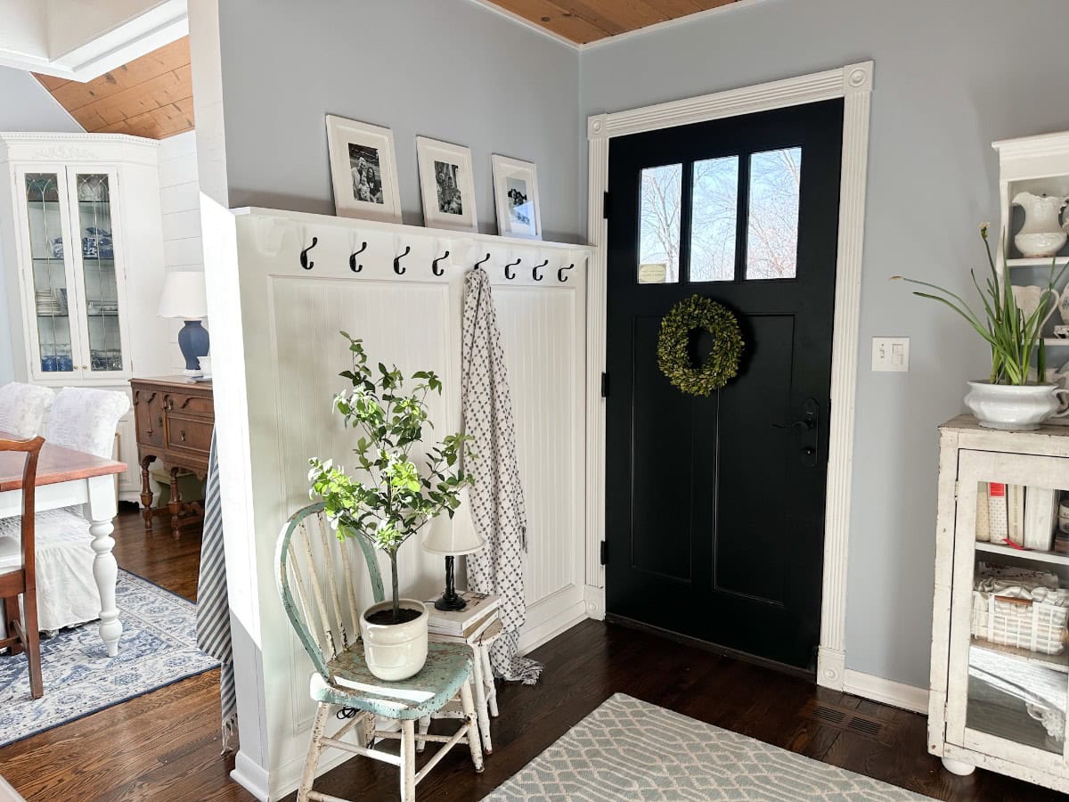Entryway with a black door adorned with a green wreath. A patterned scarf hangs on a white coat rack next to framed photos. A small plant sits on a chair, and light enters through the door's glass panes, illuminating the wooden floor.