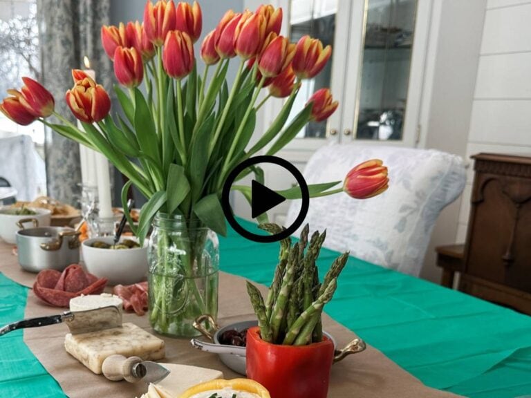 A table is set with a green tablecloth, adorned with red and yellow tulips in a jar. Various cheeses, asparagus in a red pot, and charcuterie are spread across the table. A cushioned chair and china cabinet are visible in the background.